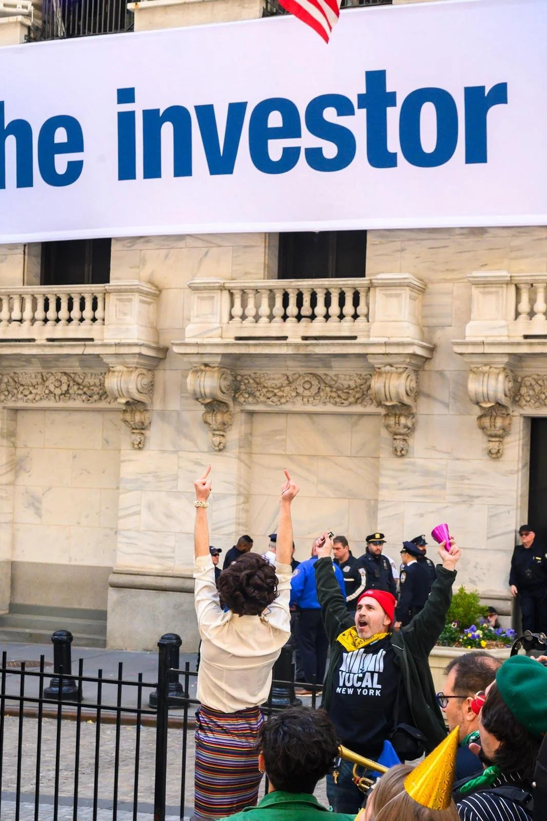 People protesting outside a building with a large sign that reads 'the investor.' Several police officers are in the background, and some protesters are wearing colorful clothing and holding signs.
