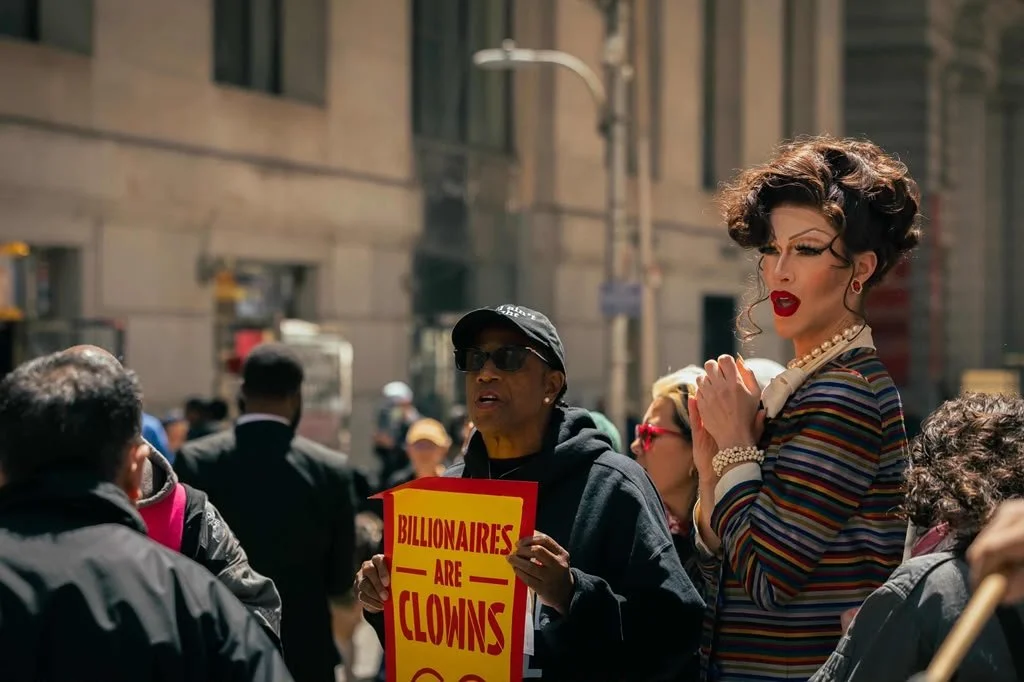 A crowd of people on a city street, with one woman holding a yellow sign that reads 'Billionaires Are Clowns.' A tall person dressed as a drag queen with makeup and a striped dress stands next to her.