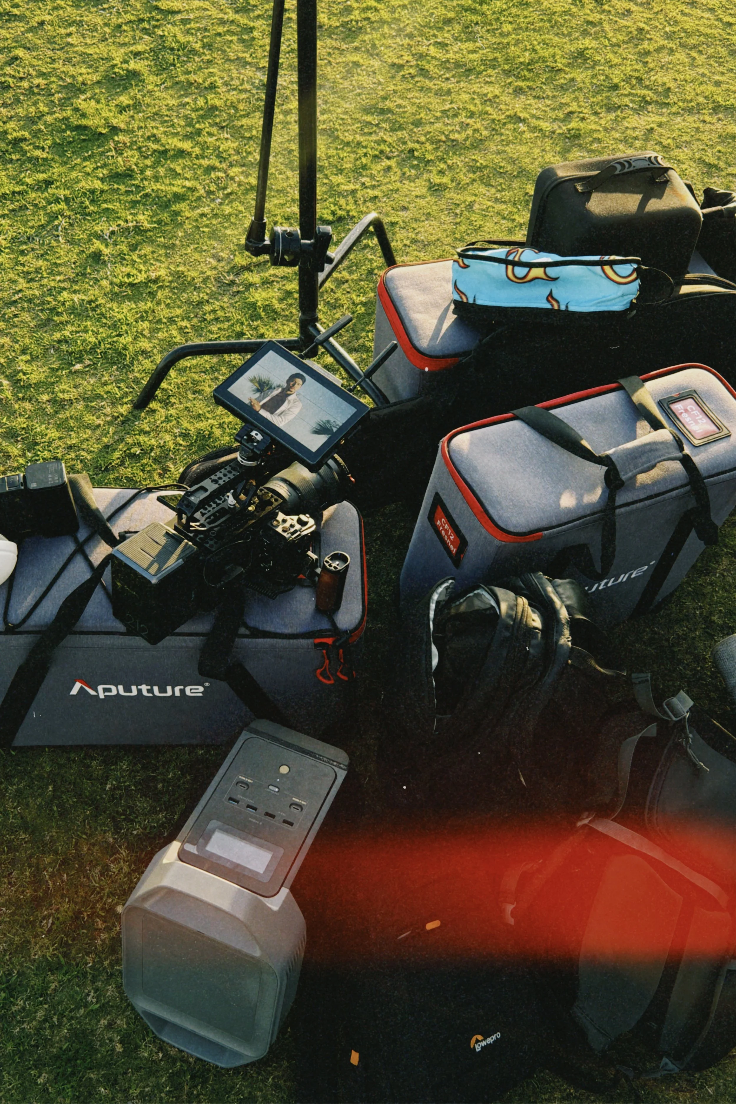 Photography equipment and camera gear laid out on grass including a camera with a monitor, a Lowepro camera bag, a speaker, a power station, and various accessories.