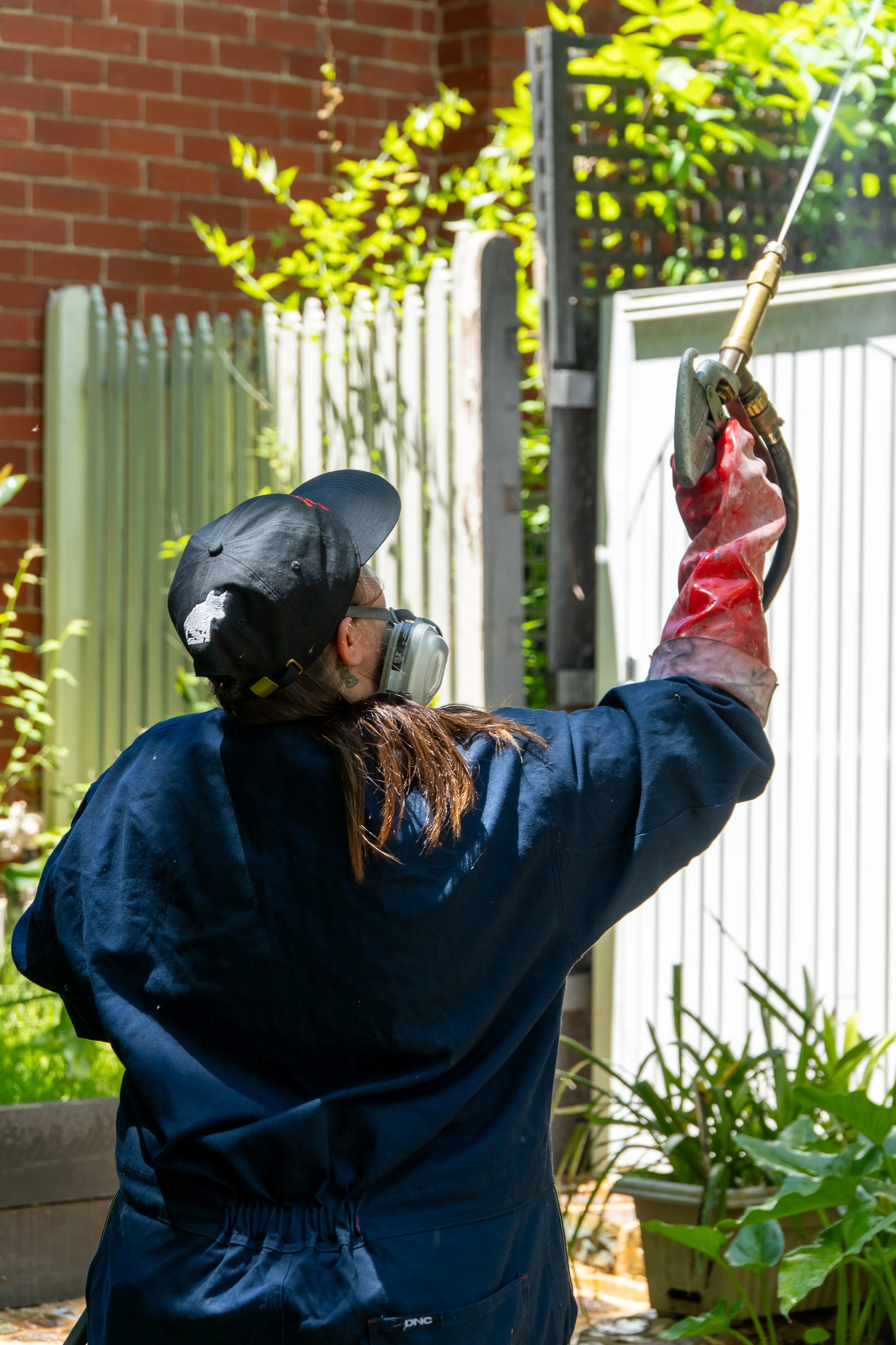 A person in a dark blue jacket, black cap, and protective mask holding a gardening tool with a red glove, watering plants in a garden with green plants and a brick wall background.