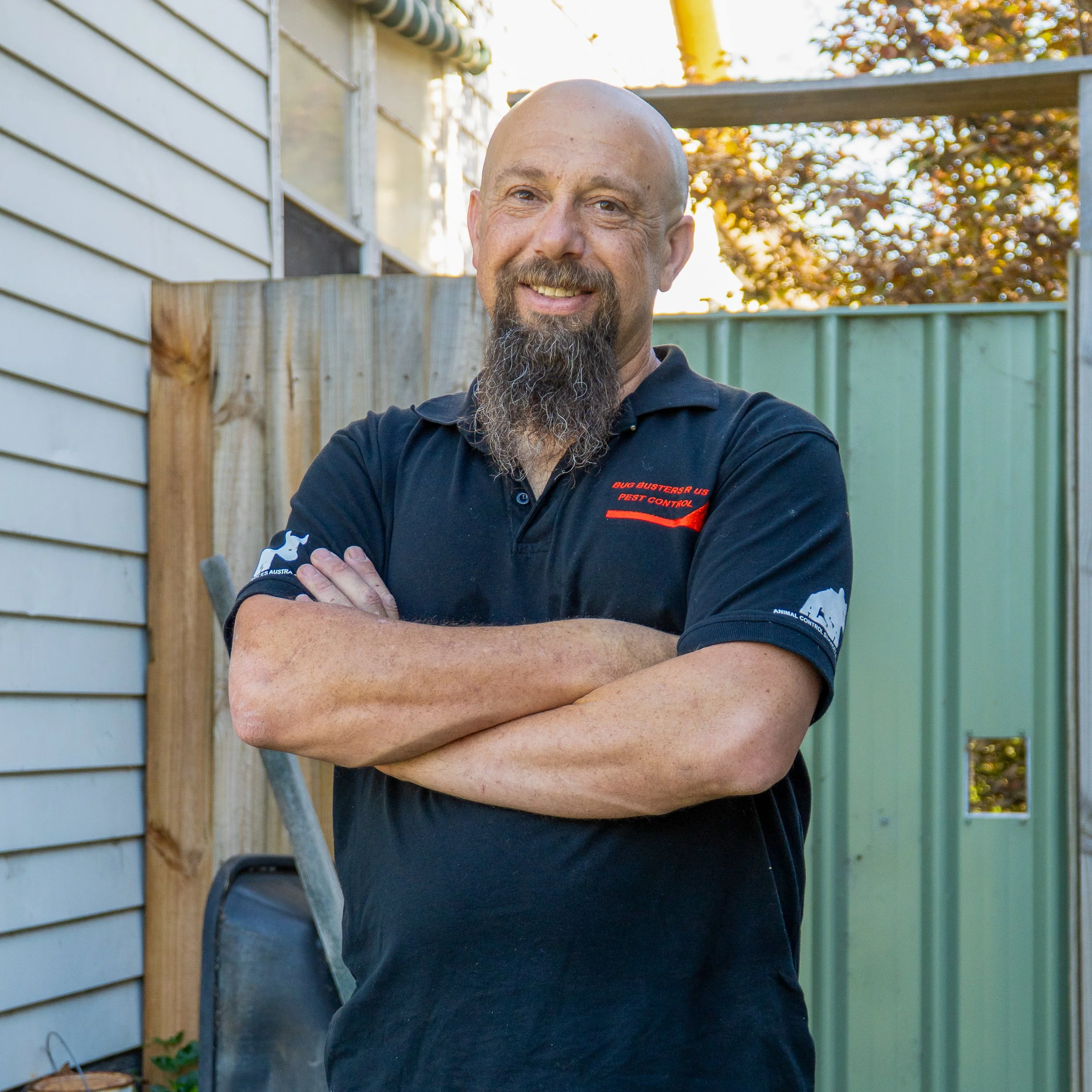 A man with a bald head, beard, and mustache smiling with arms crossed, wearing a navy blue shirt with a logo on the sleeve, standing outdoors during autumn with trees in the background and a fence on the left.