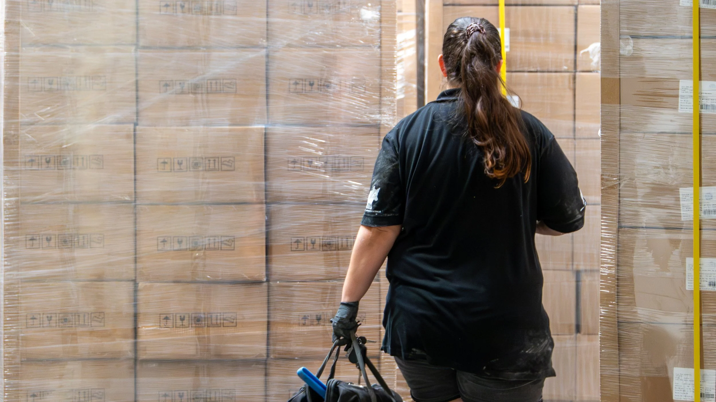 A person with brown hair tied back, wearing a black shirt and black gloves, standing in front of stacked cardboard boxes wrapped in plastic in what appears to be a warehouse or storage facility.