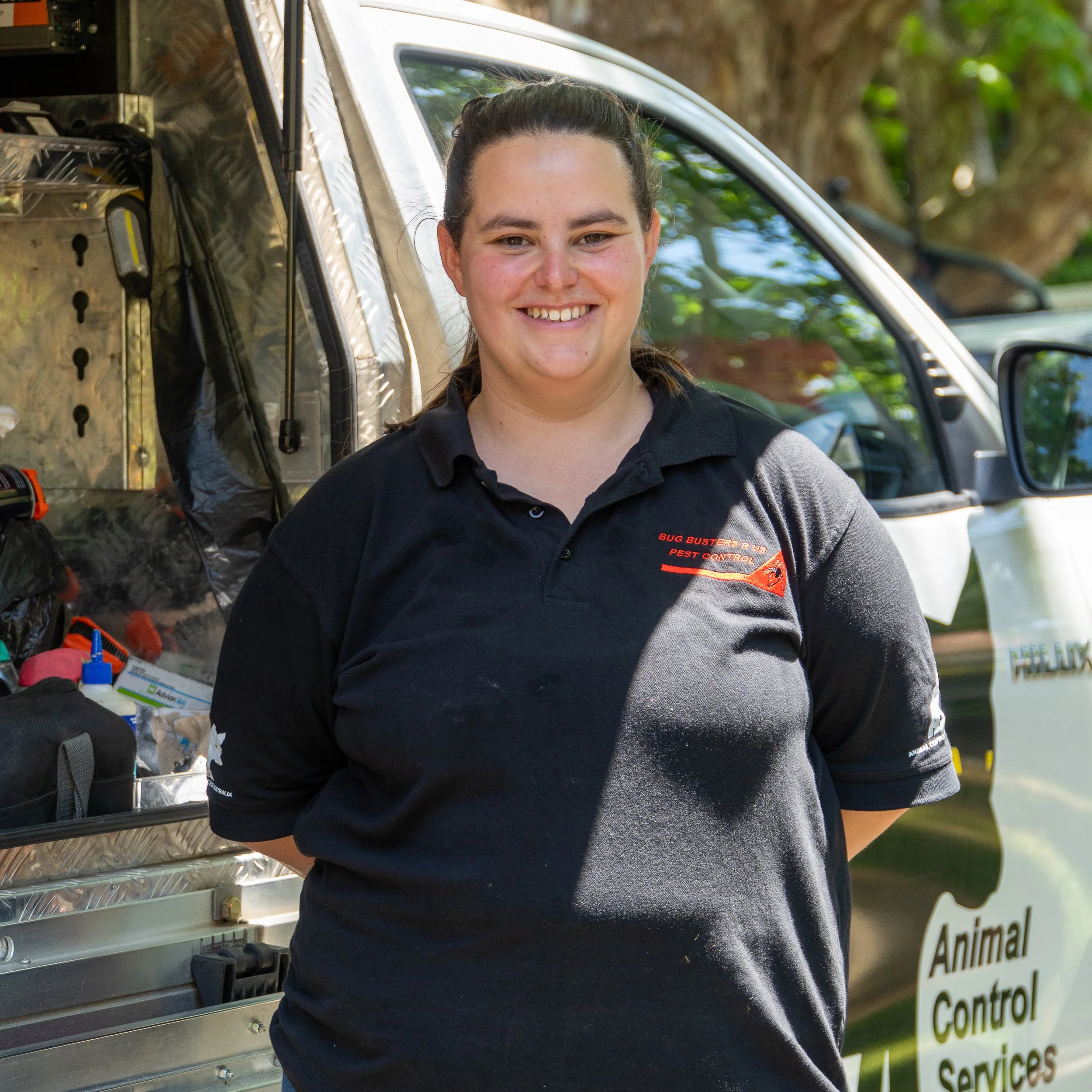A young woman with a smile and brown hair pulled back, standing outdoors next to a van with pest control equipment. She is wearing a black shirt with red and orange text that says 'Bug Busters' and 'Pest Control'.