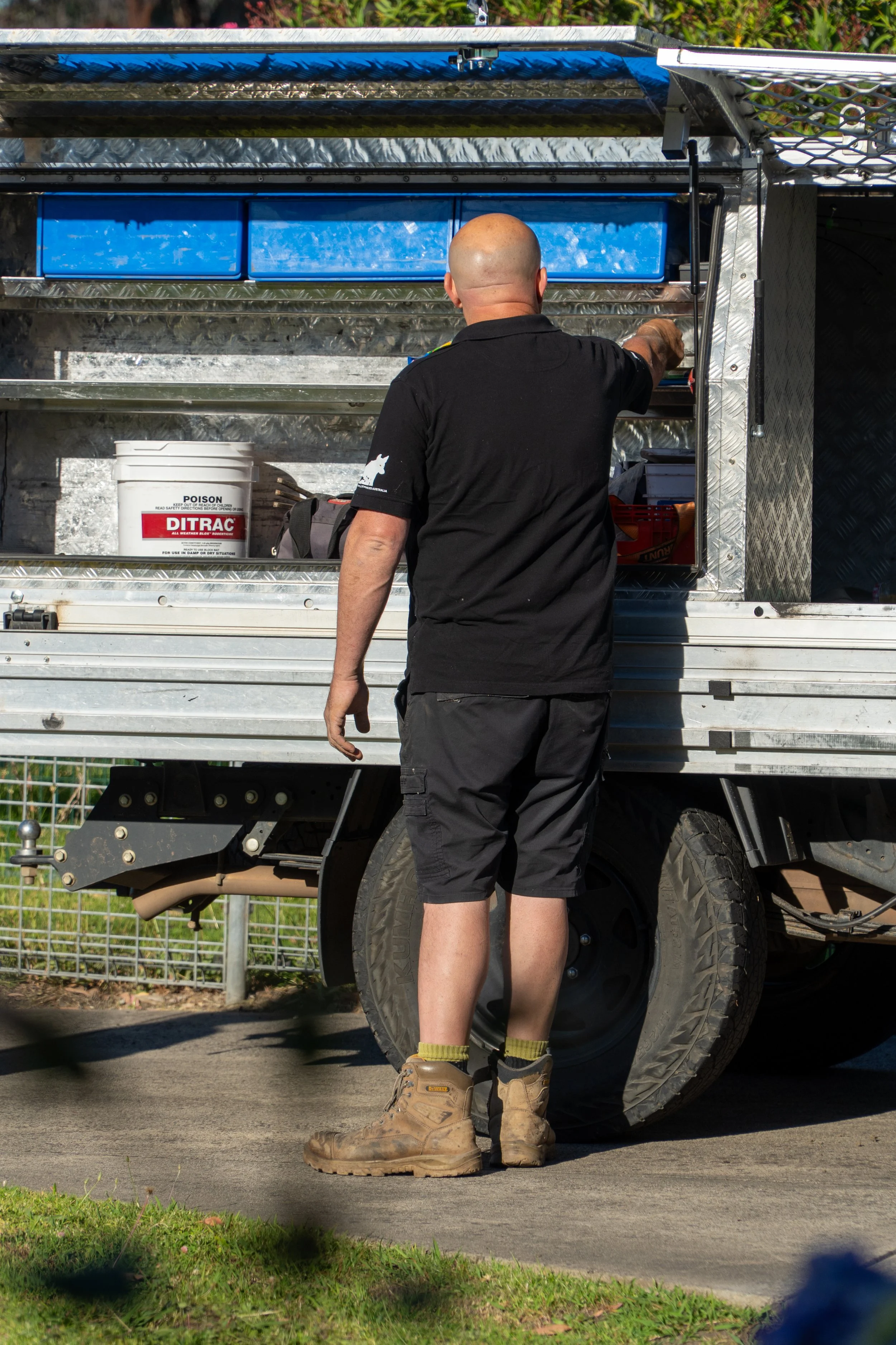 A bald man wearing a black T-shirt, shorts, and tan boots is standing in front of an emergency vehicle or trailer, reaching into it. The vehicle has a metallic exterior with toolboxes, a white bucket labeled 'DITRAC' and a green garden edging visible.