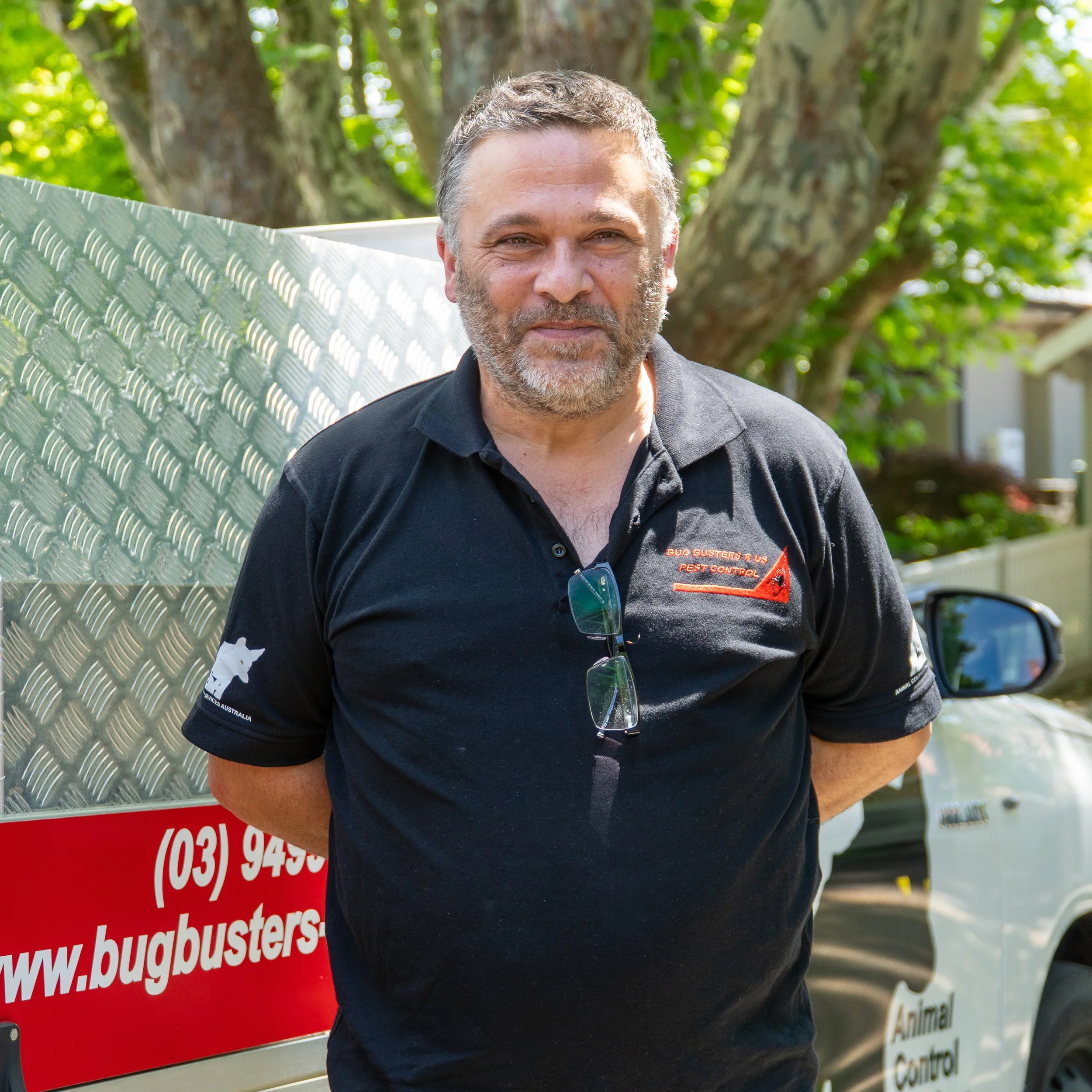 A man with short gray hair and beard wearing a black Polo shirt with orange and white text, standing outdoors in front of a large tree with green foliage. He is smiling slightly and has sunglasses hanging from his shirt collar. Part of a red and white sign with a website including the text "bugbusters" is visible in the foreground.