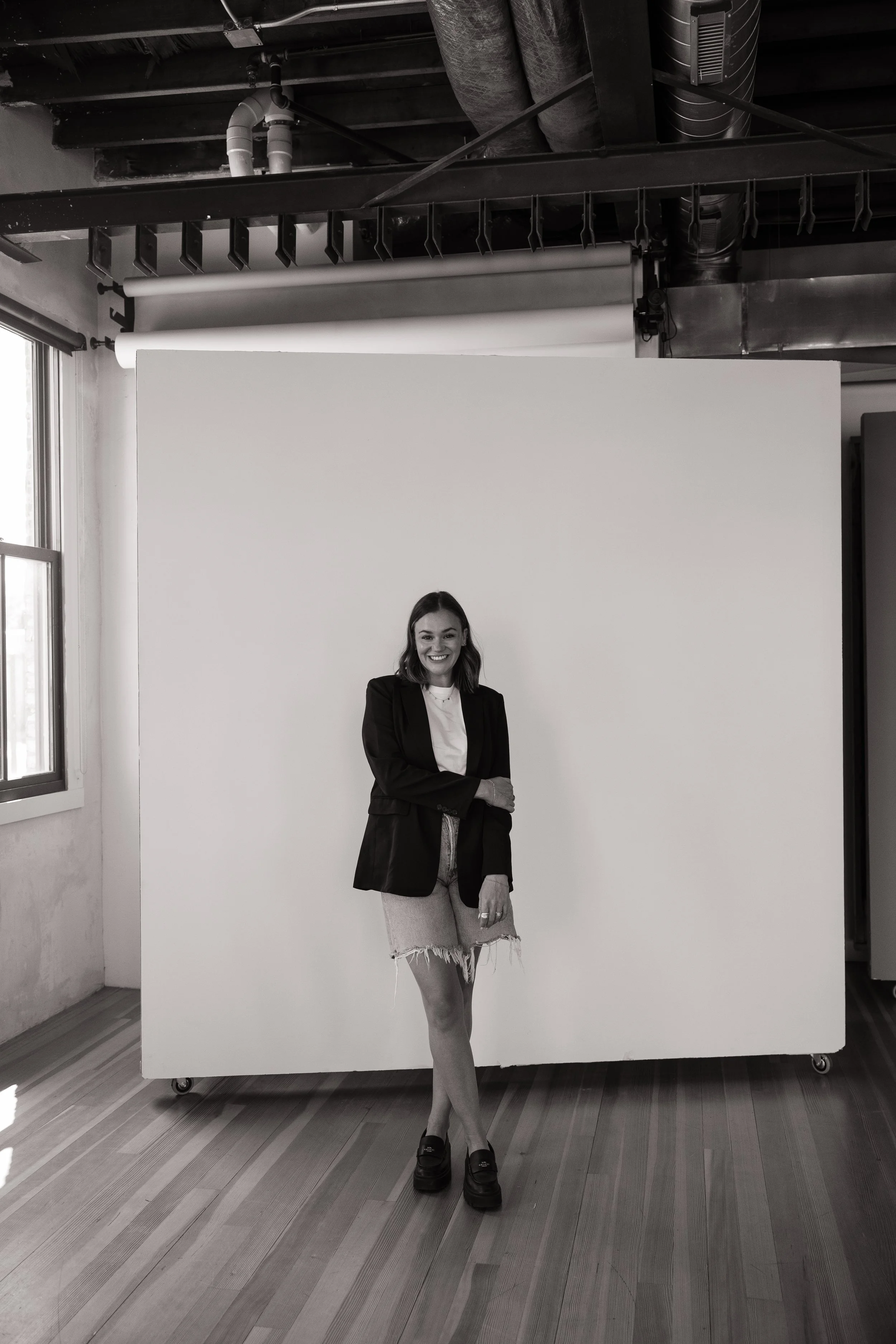 A woman with long hair standing in front of a large white backdrop in a photography studio, wearing a blazer, ripped shorts, and platform shoes, smiling at the camera.
