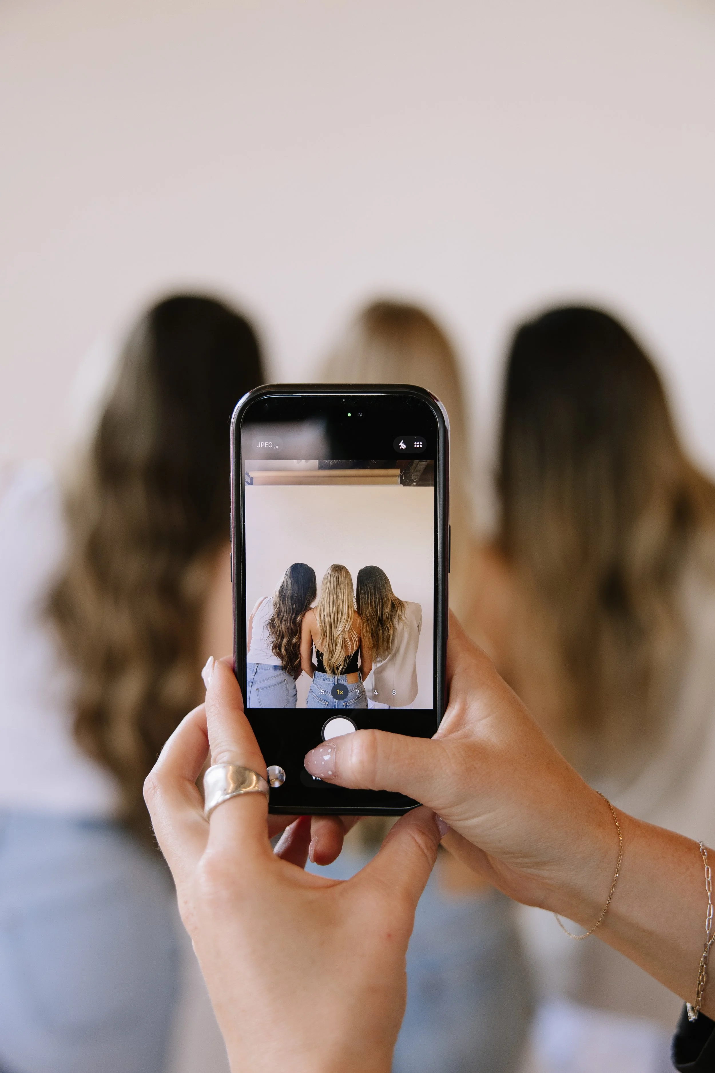 Person holding a smartphone taking a photo of three women with long hair, standing together, viewed on the phone's screen.