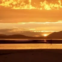 Sunset over a body of water with mountains in the background and two people walking along the shore.