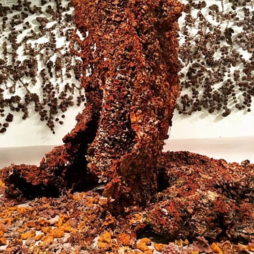 A twisted, rust-colored sculpture resembling a tree trunk, made of textured, rough material, with small rocks or gravel at the base and a background decorated with numerous dark, rounded objects on a white surface.