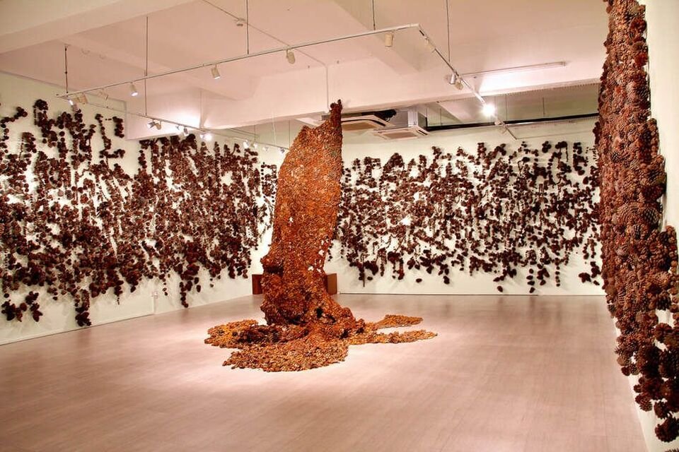 An art installation consisting of numerous pinecones attached to white walls, with a large, vertical, twisting structure covered in pinecones in the center of the room.
