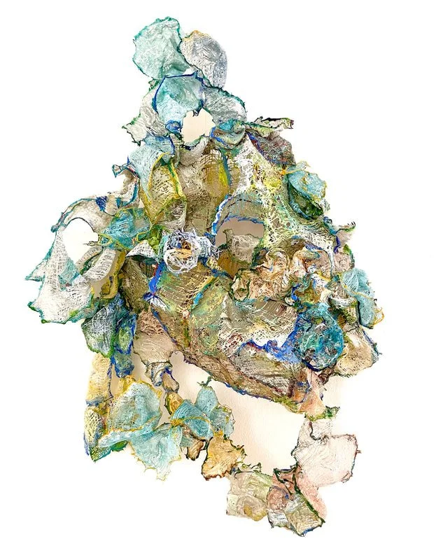 An abstract artwork resembling a human figure, composed of textured and colorful fabric scraps with shades of blue, green, yellow, and beige.