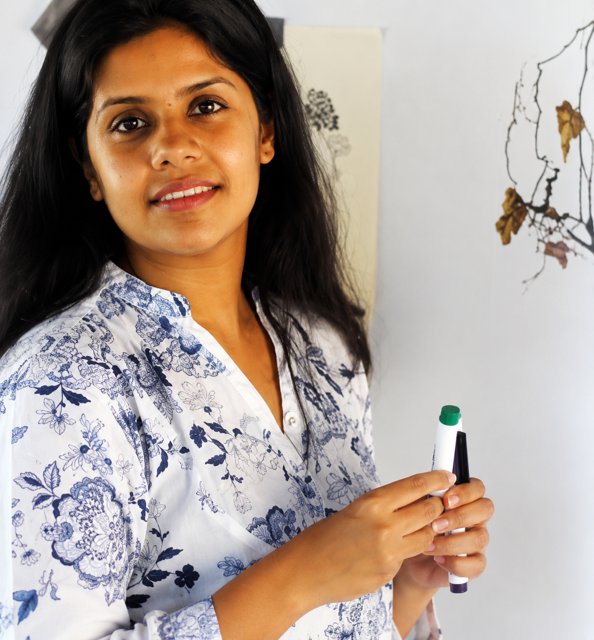 A woman with long black hair holding a whiteboard marker, standing in front of a whiteboard with some drawings, wearing a white blouse with blue floral patterns.