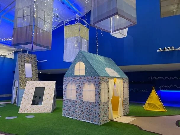 Indoor play area with a fabric playhouse and small yellow tent on artificial grass floor under colorful hanging decorations.