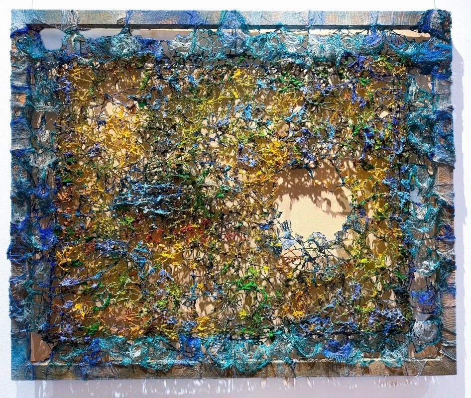 Colorful abstract textile art with a circular opening, featuring blue, yellow, green, and black threads in a tangled pattern, bordered with fabric strips.