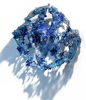 A complex, irregularly shaped blue glass sculpture with reflective qualities, casting shadows on a light background.
