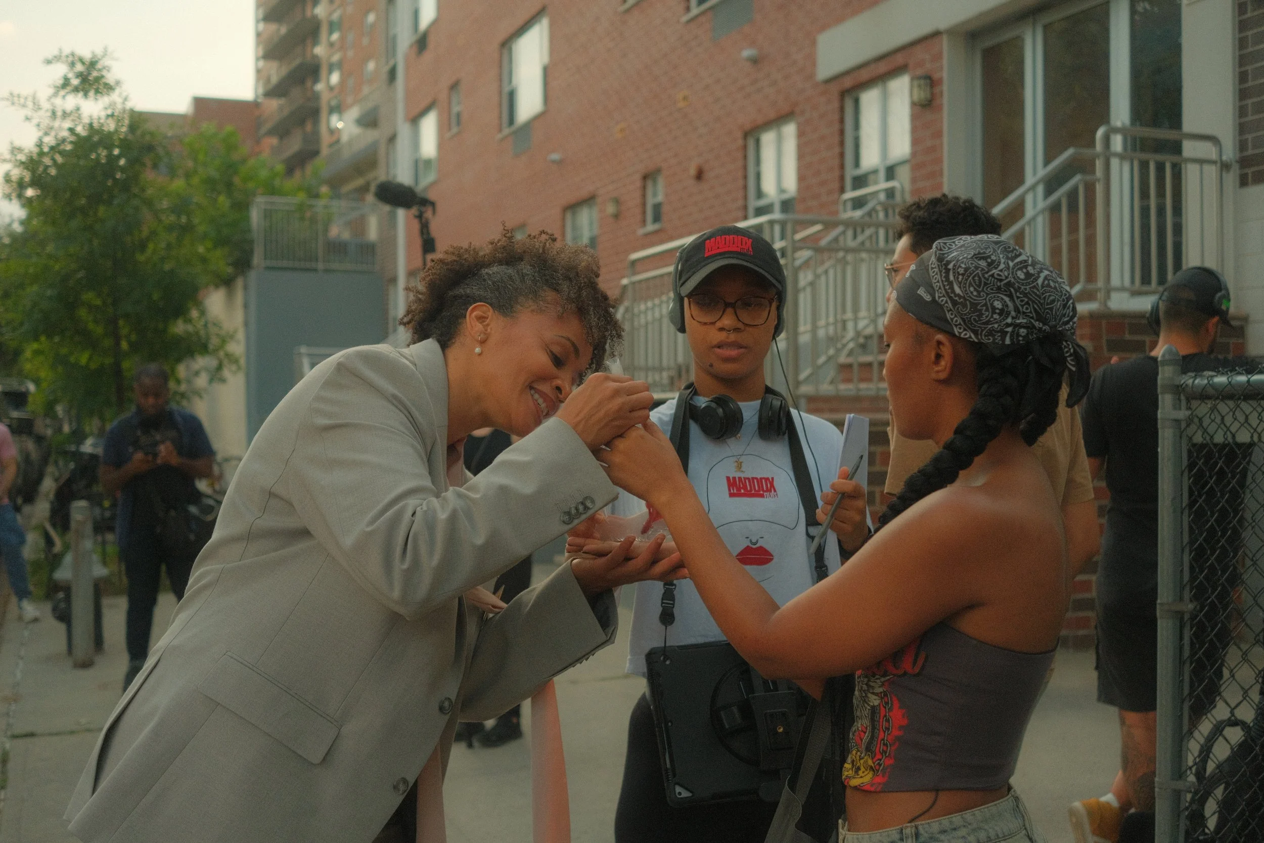 A person in a light gray blazer smiling as they hold hands with a topless man wearing a bandana and colorful shorts, standing behind a chain-link fence. Around them, a group of people are on the street near brick apartment buildings, with some holding cameras and equipment.