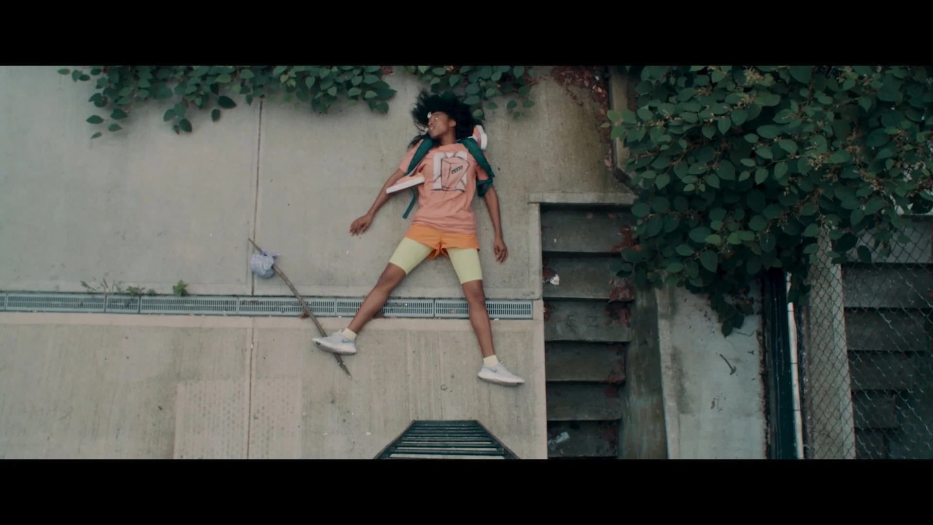 A girl lying on her back on a concrete sidewalk, smiling, with green bushes and a fence behind her, wearing a peach T-shirt with a juice box graphic, yellow shorts, white sneakers, and a backpack.