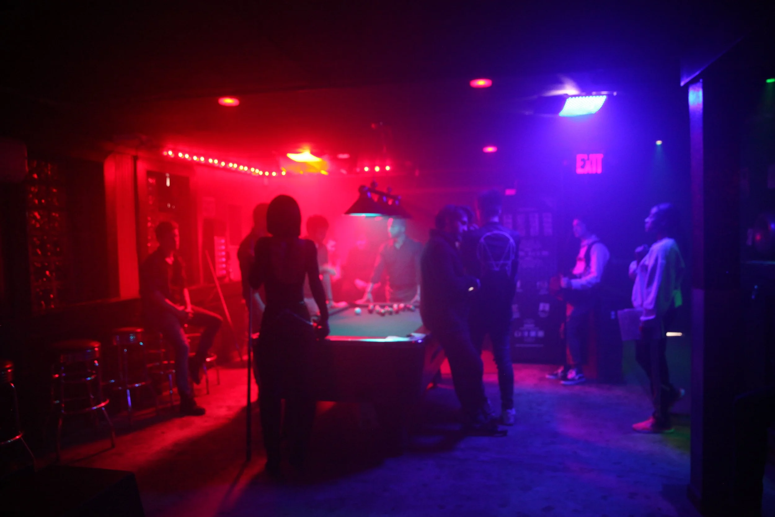 People gather in a dimly lit nightclub area with red and blue lighting, standing around a pool table in short film Eight