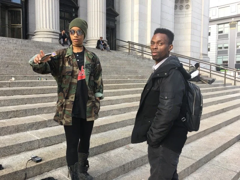 Two young adults standing on the steps of a large building with columns, possibly a government or institutional building, with other people in the background. The woman on the left is wearing a camouflage jacket, beanie, sunglasses, and black boots, 