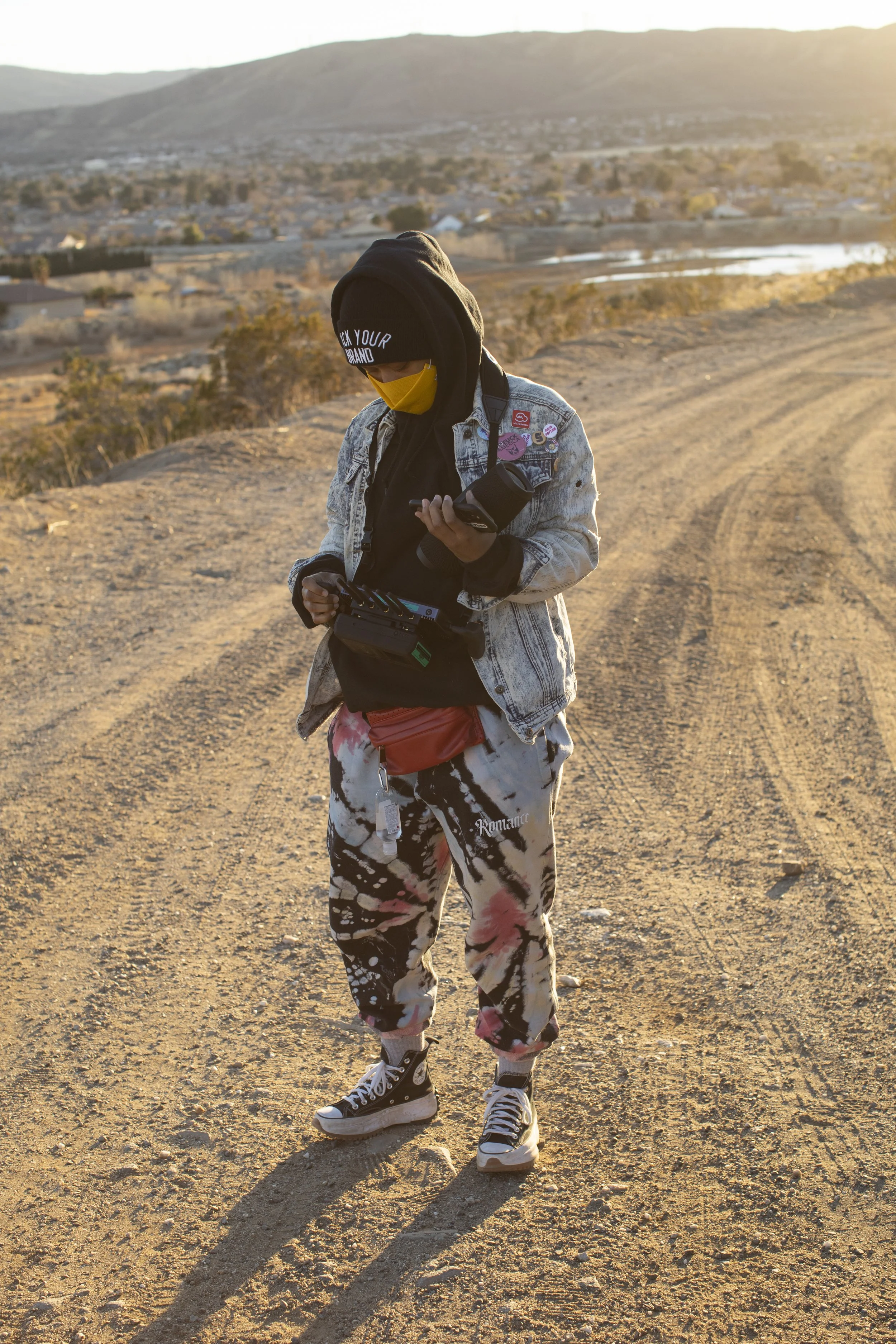 Person standing outdoors on a dirt path during sunset or sunrise, wearing a black beanie, yellow face mask, denim jacket, patterned pants, and sneakers, holding a camera and phone, with a hilly landscape and water bodies in the background.