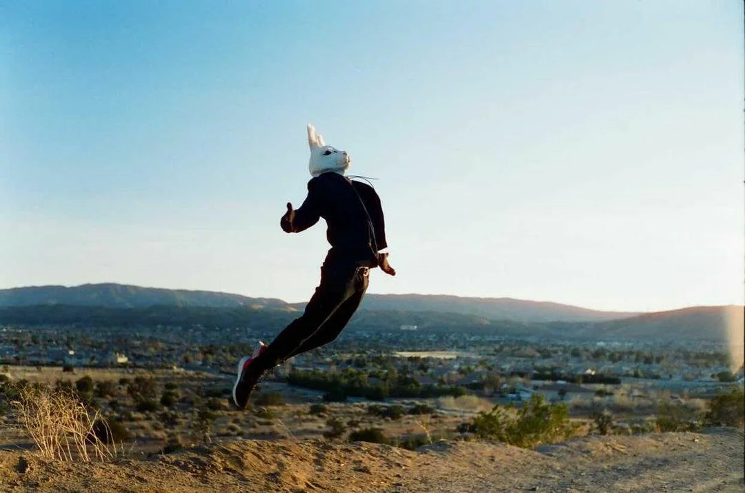 Person wearing a rabbit mask jumping in mid-air against a desert landscape with mountains in the background at sunset from Hailes music video Escape