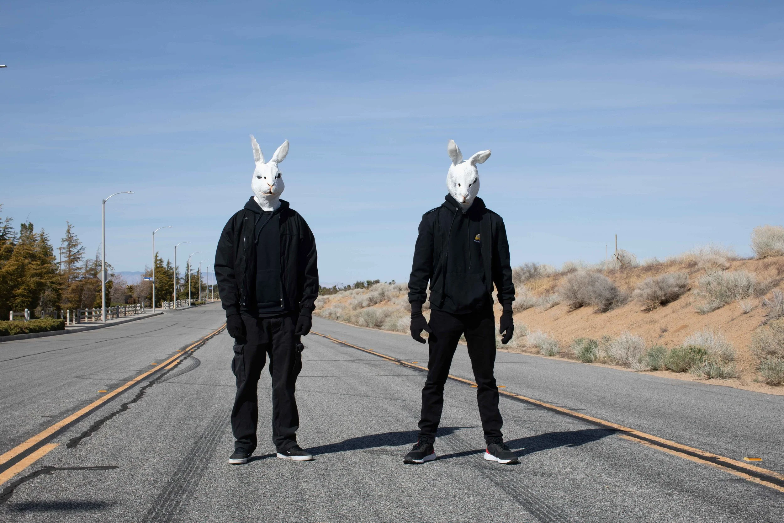 Two people walking down the middle of an empty road, wearing large white rabbit mask costumes and black hooded jackets under a clear blue sky.