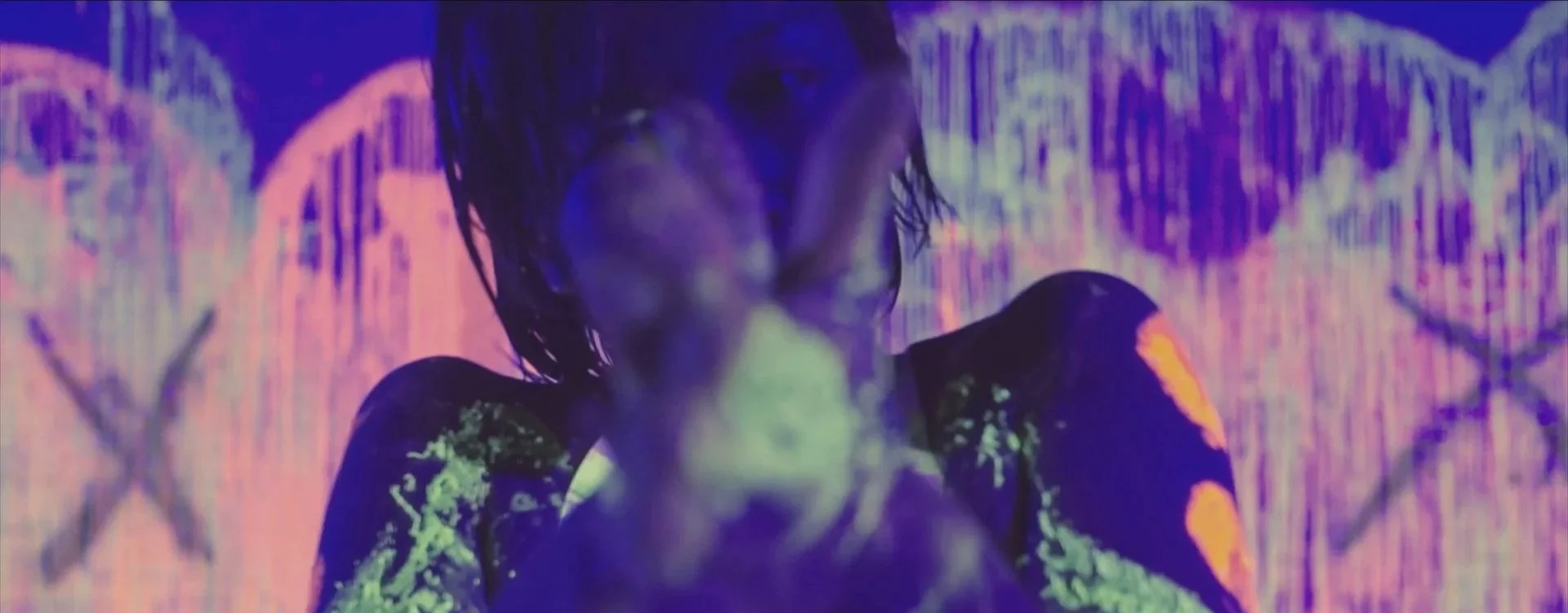 A person with wet, dark hair and dark clothing is exhaling smoke in a dimly lit setting with colorful, neon-like background visuals.