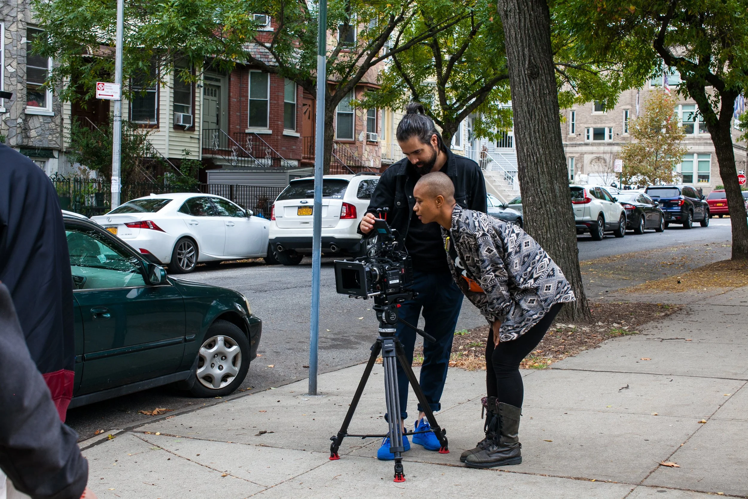 Director Louvinia and DP Erin Collette on a city sidewalk are adjusting professional filming equipment, with parked cars and residential buildings in the background.