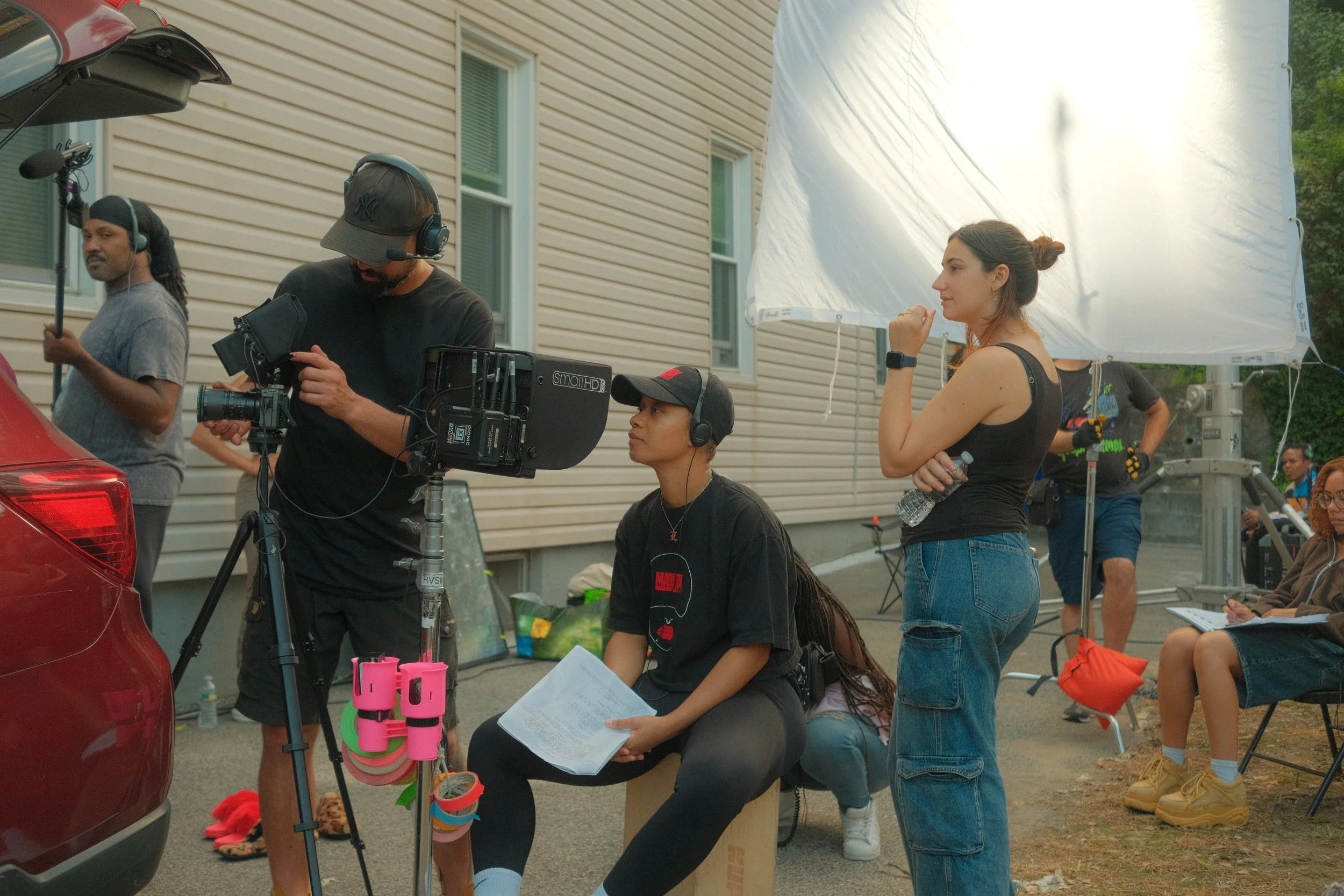 Betta Fish film crew setting up equipment outdoors, including camera, lights, and reflectors, as they prepare for a shoot near a residential building during the daytime.