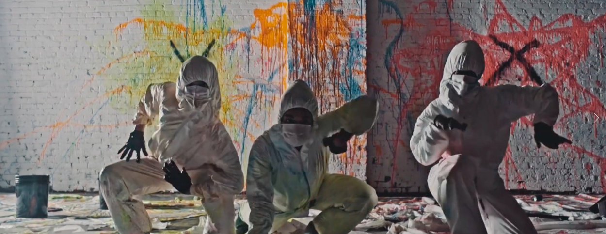 Three individuals dressed in white protective suits, masks, and gloves are crouched down in front of a graffiti-covered wall with splashes of colorful paint. They are holding spray paint cans and a paint roller.