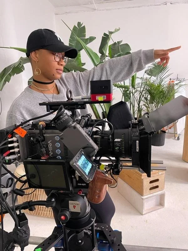 Director Louvinia wearing a cap, glasses, and jewelry pointing to the right, standing behind professional camera equipment and monitors, with large green potted plants in the background.
