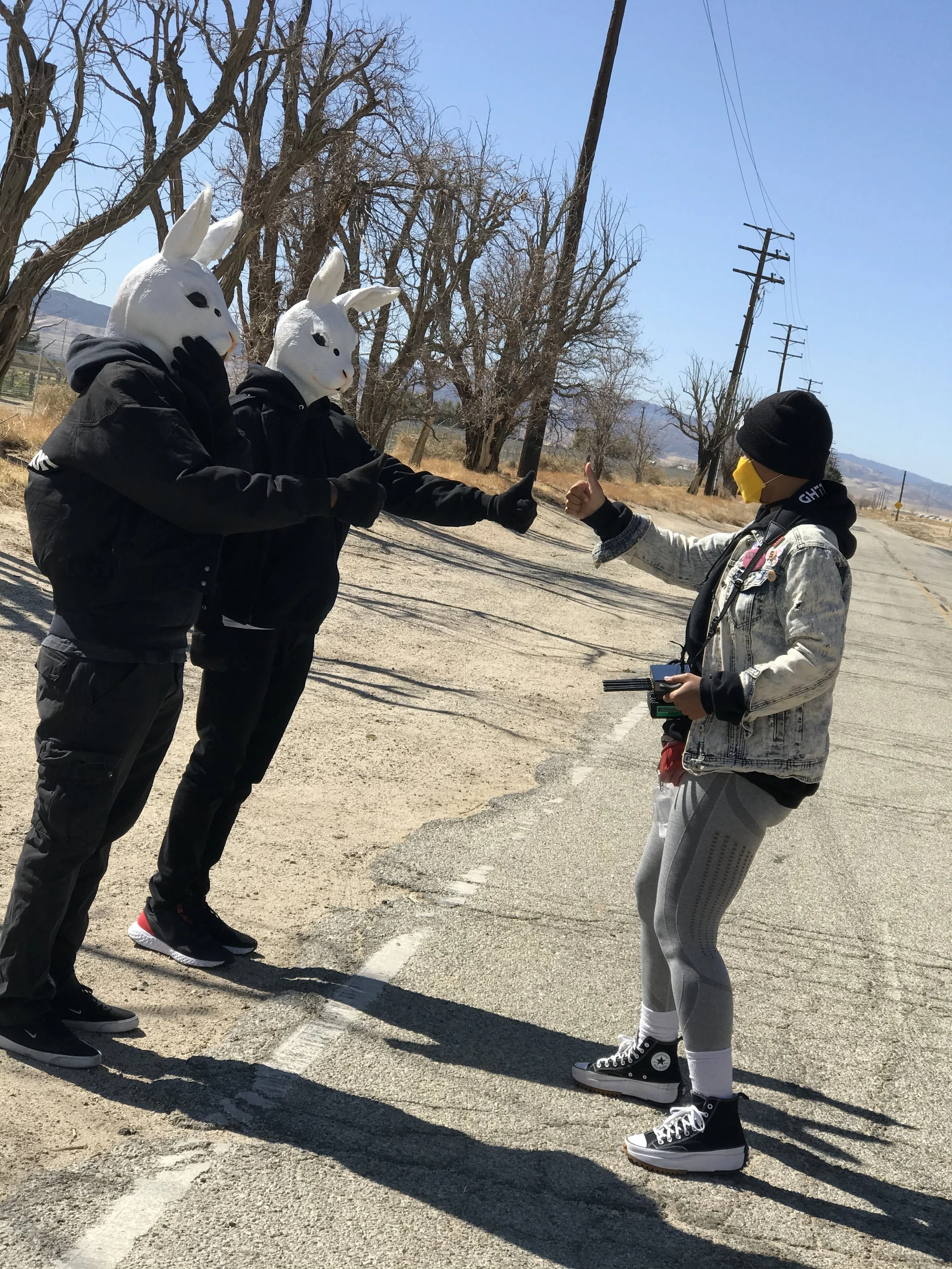 Two people wearing white bunny masks giving a thumbs-up to a person with a yellow face mask, standing in an outdoor rural area with leafless trees and mountains in the background.