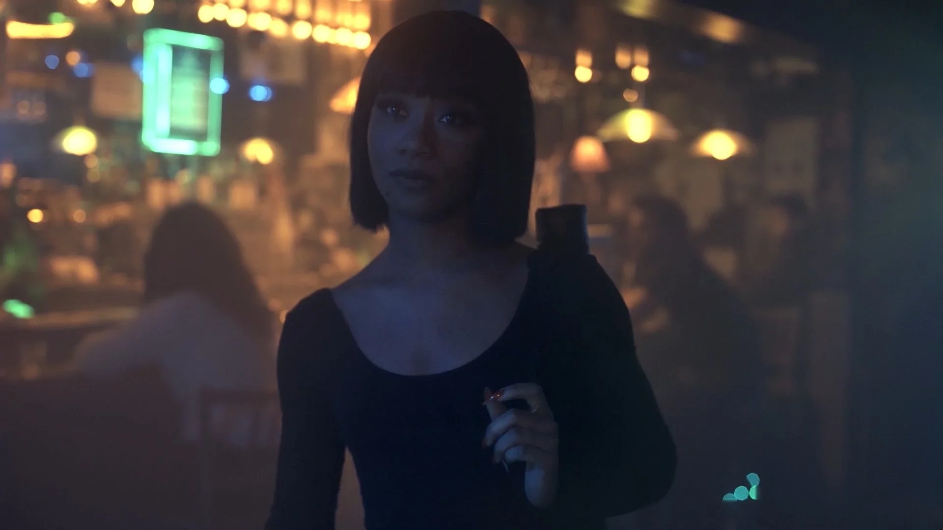 A woman with a bob haircut and dark clothing holding a cigarette in a dimly lit bar or lounge with blurred colorful neon lights in the background.