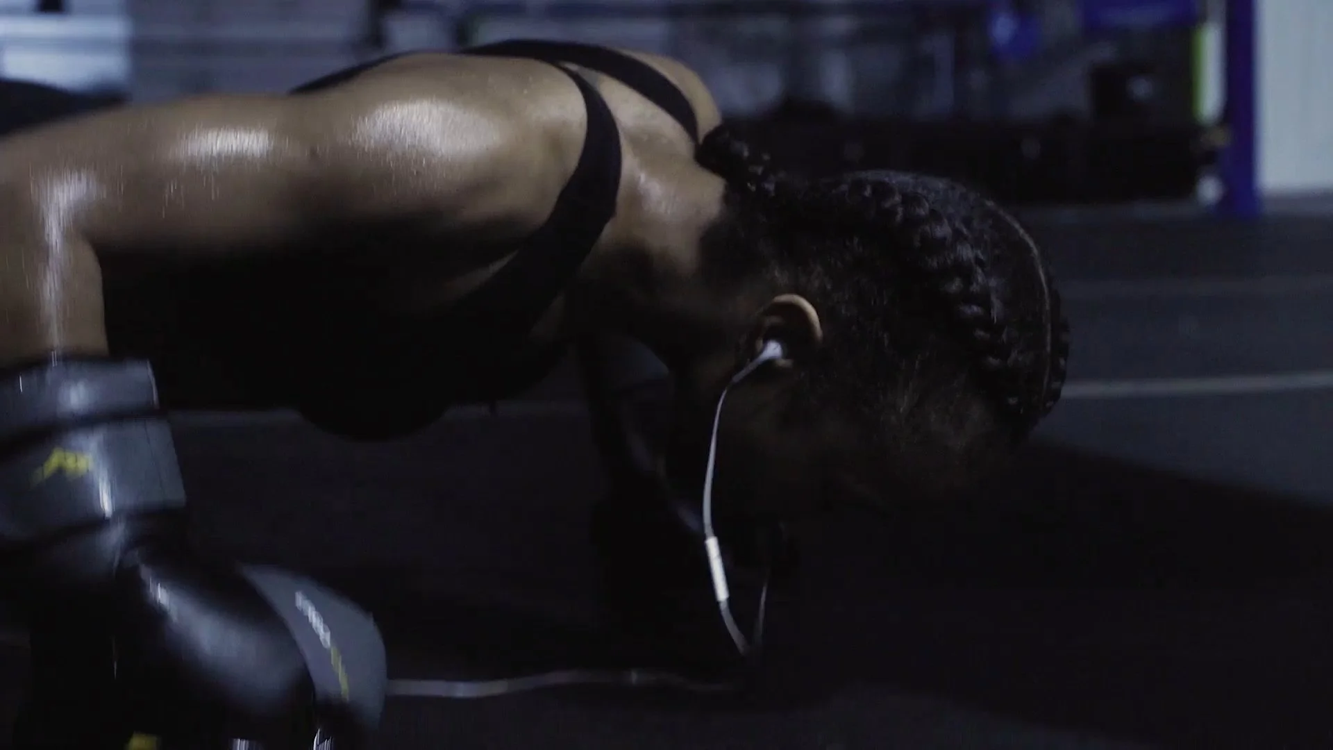 Close-up of Donna doing bush ups , wearing a black bra, with headphones in her ear, in a dimly lit room in Last Round