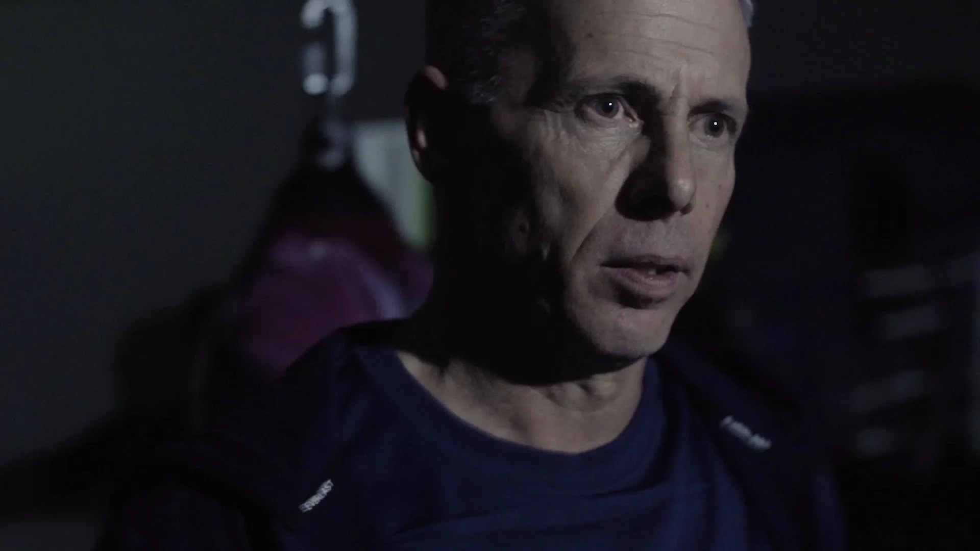 A man with short hair and light skin, wearing a dark shirt, is in a dimly lit setting, looking intently to his right with a serious expression in short film Last Round