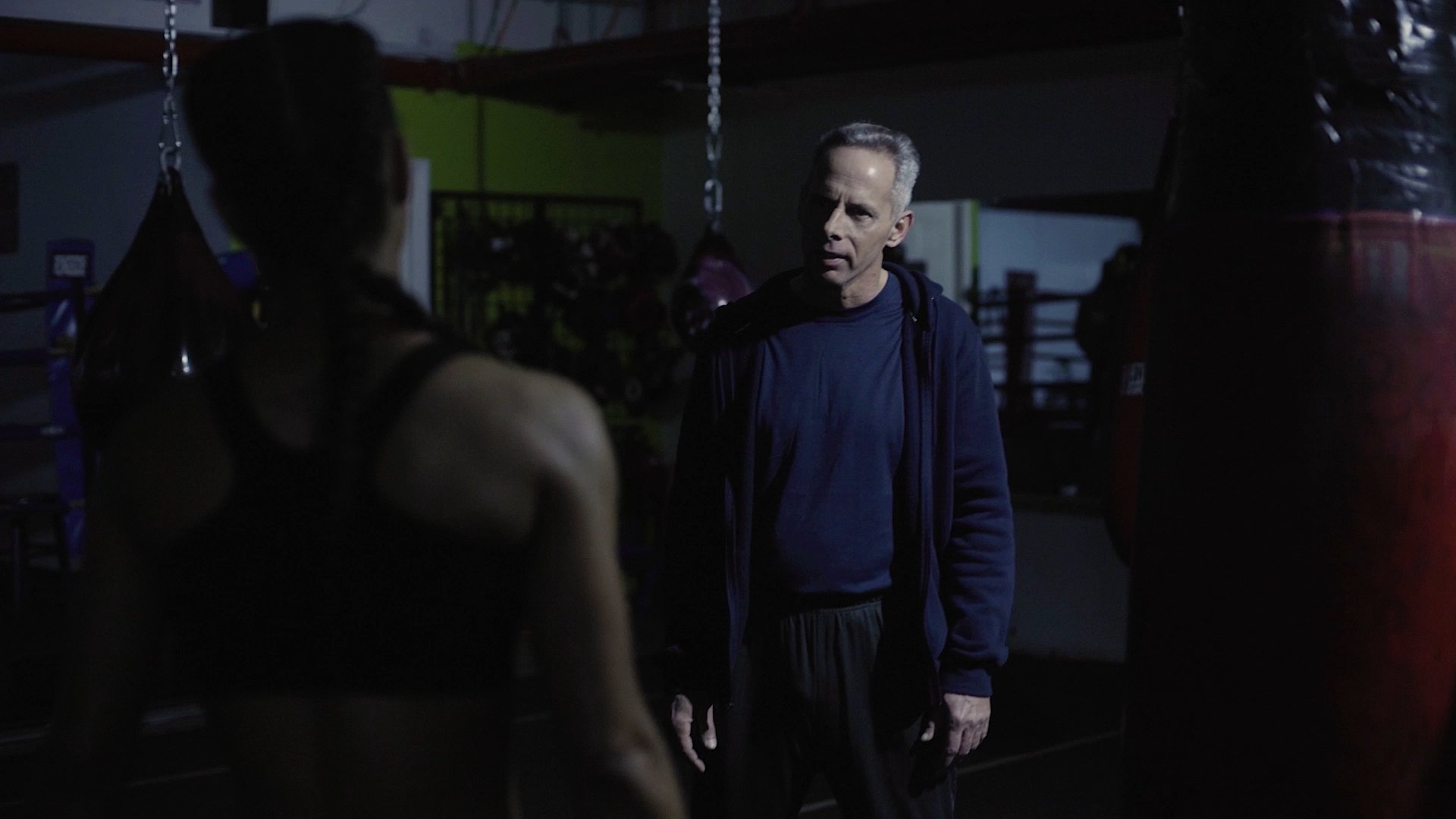 Coach with gray hair and dark clothing standing in a dimly lit gym or boxing gym, facing Donna with a sleeveless top and high braids, with punching bags and gym equipment in the background in Last Round
