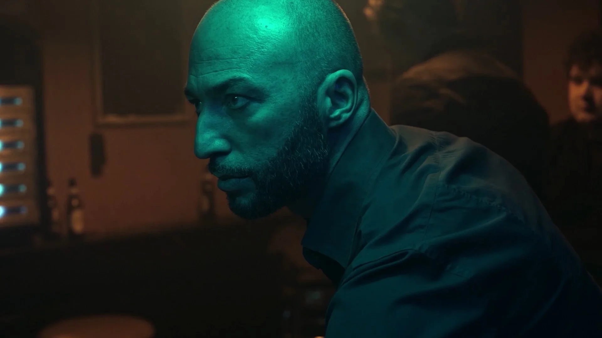 A man with a beard and a shaved head in a dark room, illuminated by teal lighting, looking to the left and sitting near audio equipment.