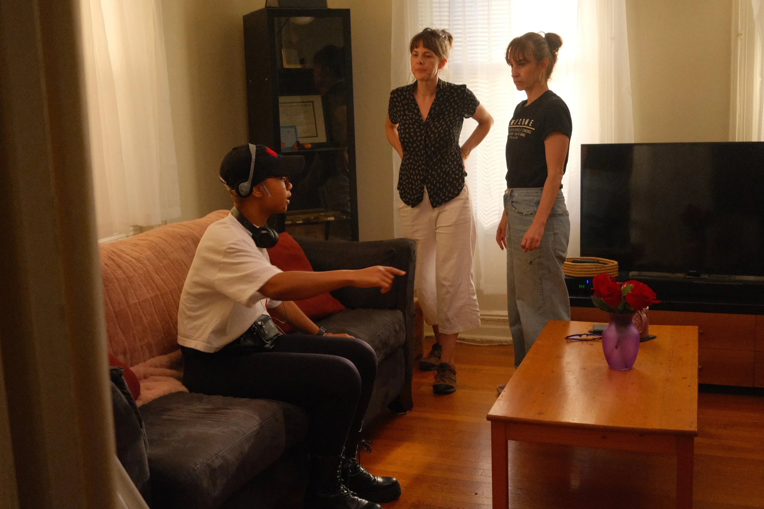 Four people in a living room. One person seated on a brown couch wearing headphones, a hat, and glasses. Two women standing near the couch, one with hands on her hips, the other looking down. A person's reflection is visible in a glass cabinet. A TV and a purple vase with red flowers on a wooden table are also in the room.