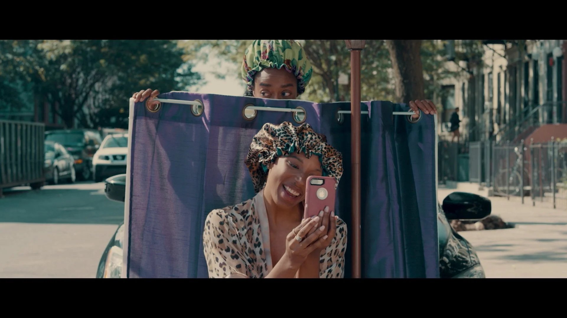Two women in leopard print clothing and matching headscarves looking and smiling at a phone, positioned inside a fabric-covered cart with purple sides, on a city street with parked cars and trees in the background.