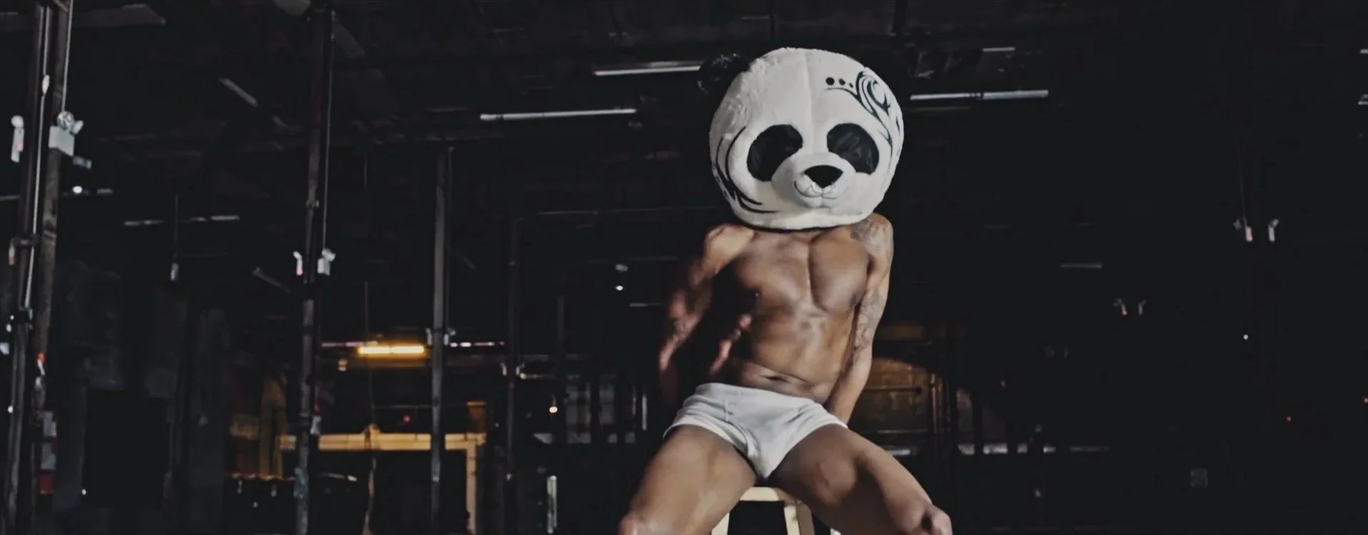 A muscular man wearing only shorts and a panda head mask sitting in a dark, industrial-like space from Cereal Killers This video