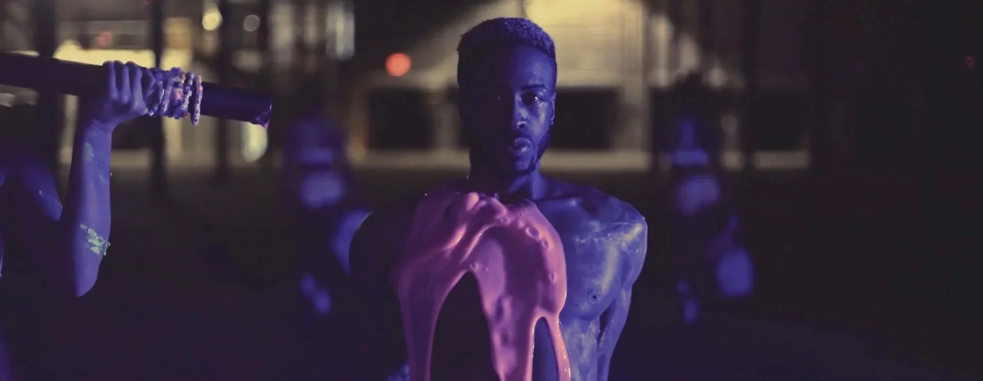 A person is sitting in a dark environment illuminated by purple and yellow lights. There is a large, pinkish-orange melting substance on their chest and shoulder, creating a surreal visual effect.