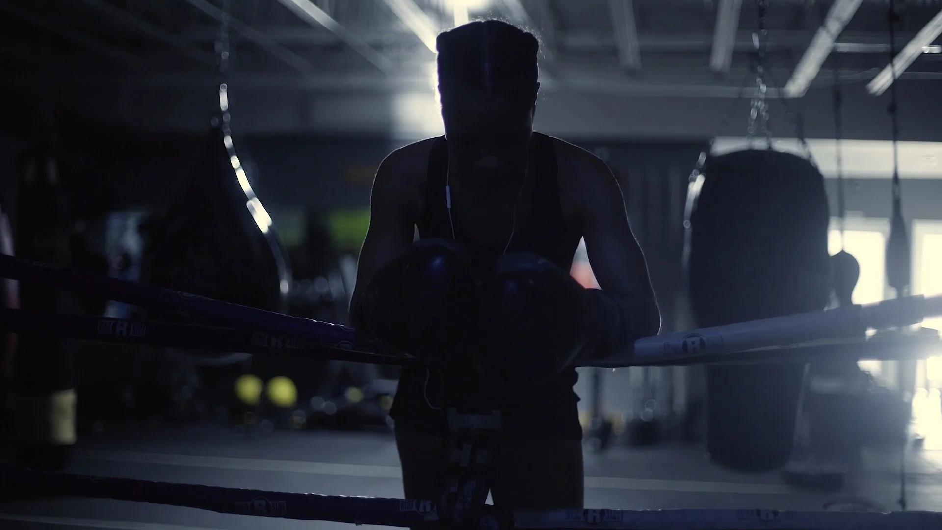 Donna in a dark gym holding a medicine ball with boxing gloves hanging on the side, with ropes and gym equipment in the background, illuminated by bright light from above in Last Round