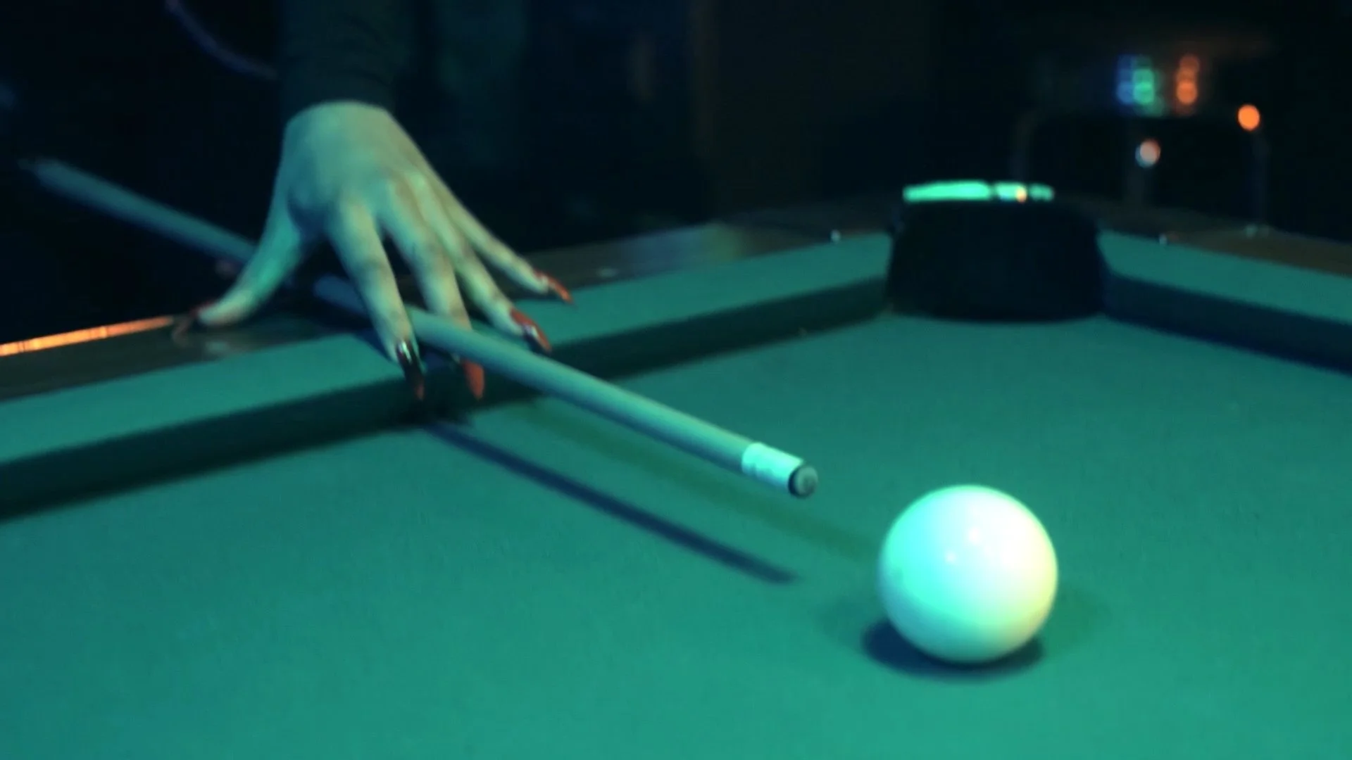 A person's hand is seen reaching over a pool table, holding a cue stick to hit a white cue ball. The scene is dimly lit, with some electronic equipment and lights visible in the background.