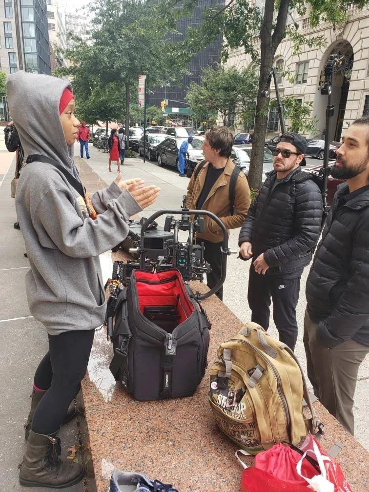A young woman in a gray hoodie and leggings, speaking to three men on a city sidewalk with filming equipment, bags, and a busy street in the background.