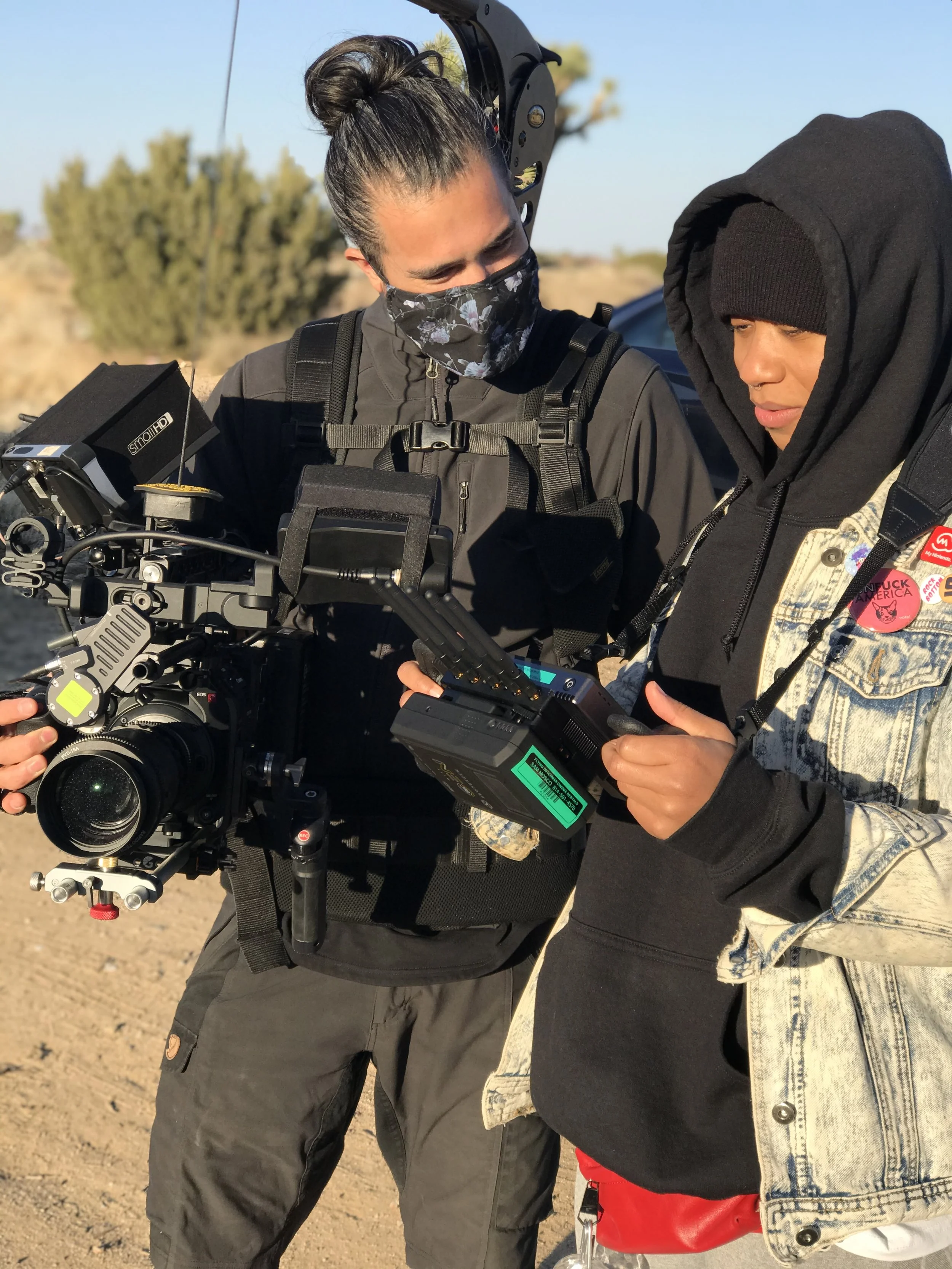 Director Louvinia and Erin Collette preparing to shoot in the desert. Erin is holding a professional camera with a large lens and accessories, wearing black jacket and face mask, Louvinia  is wearing a black beanie, hoodie, mask and holding a monitor