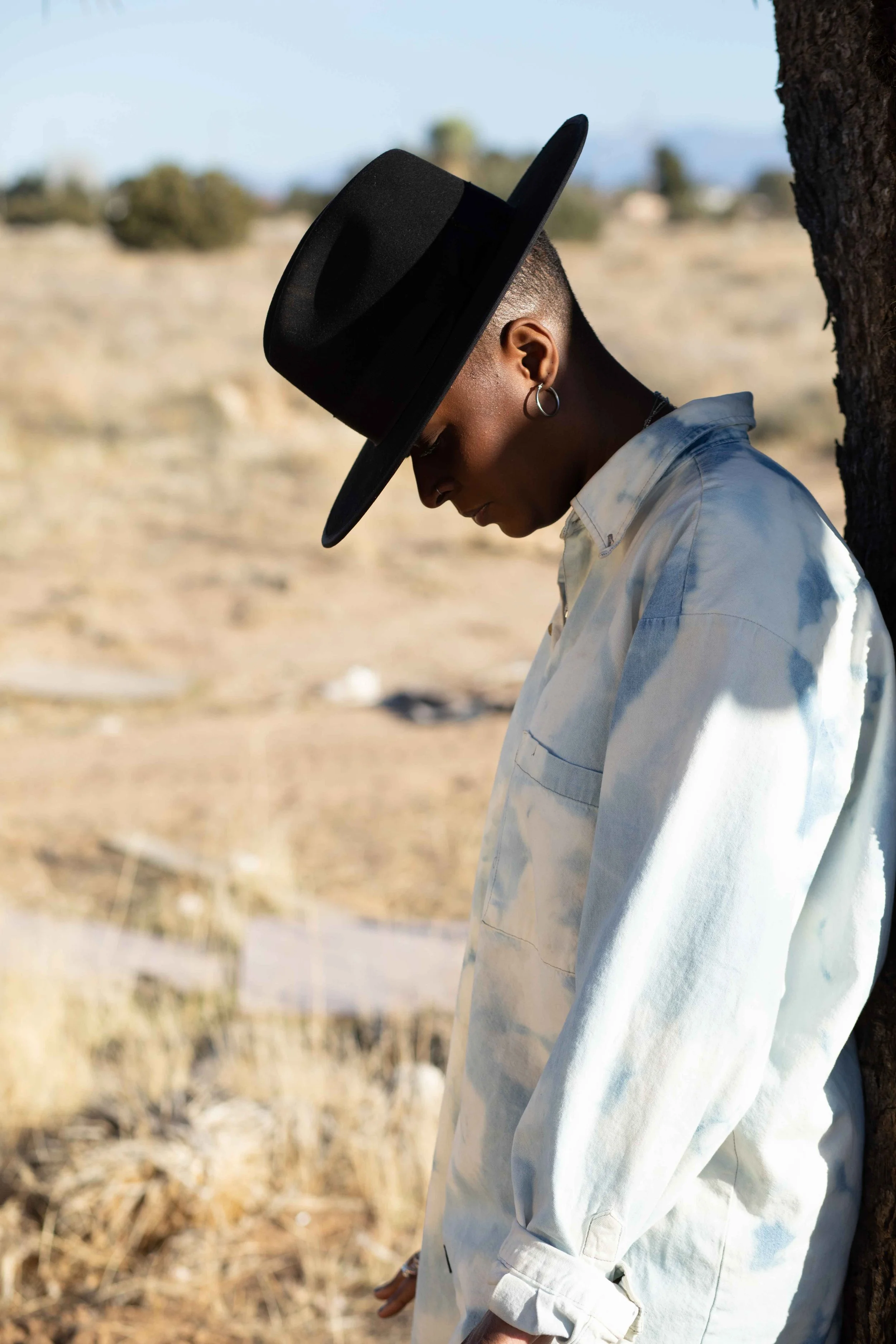 Hailes leaning against a tree, wearing a wide-brimmed black hat, a light-colored button-up shirt with sleeves rolled up, and standing outdoors in a dry, desert-like landscape with blurred background of bushes and mountains in Escape