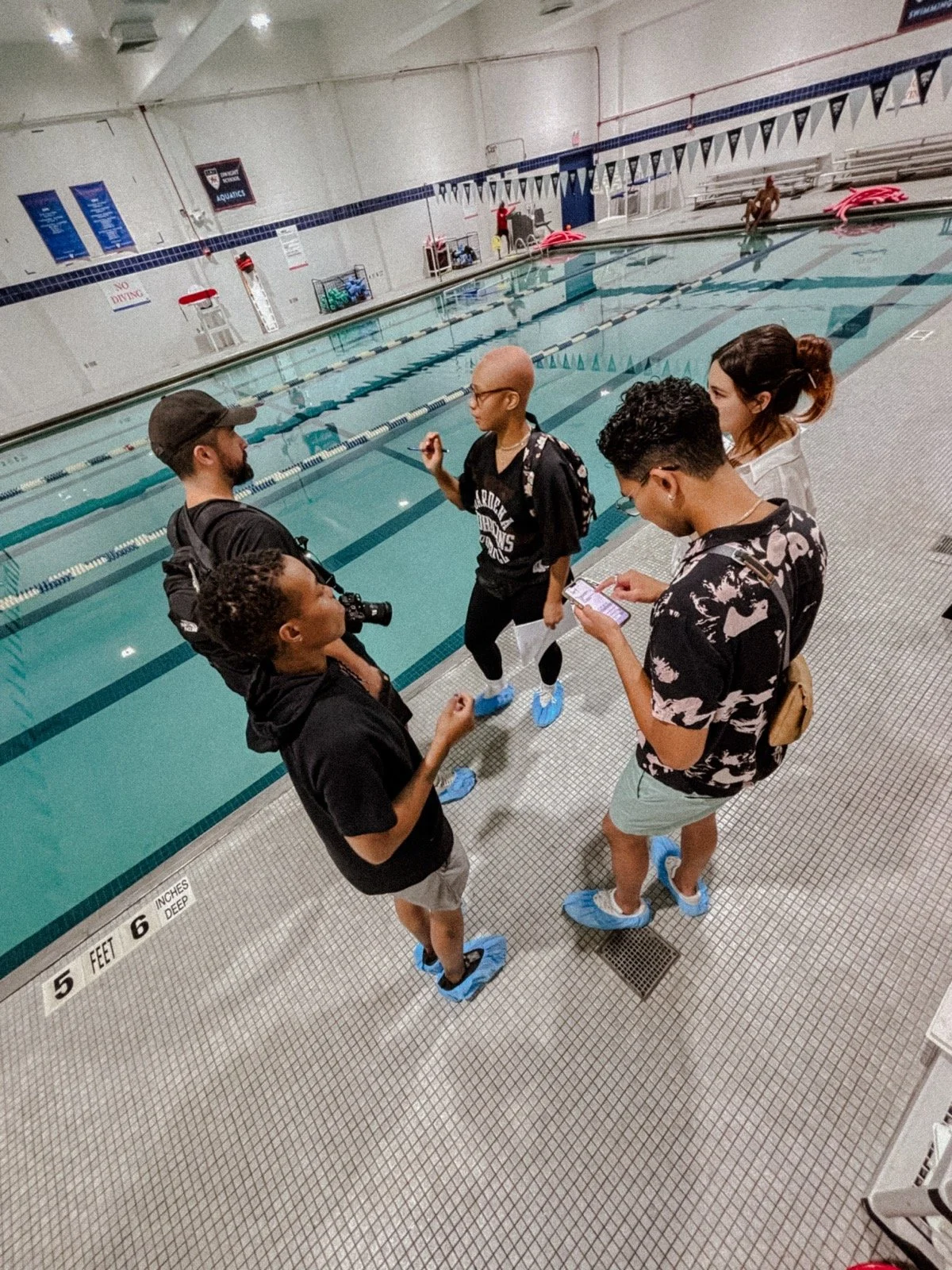 Group of five young people wearing shoe covers and face masks at an indoor swimming pool, engaged in conversation.