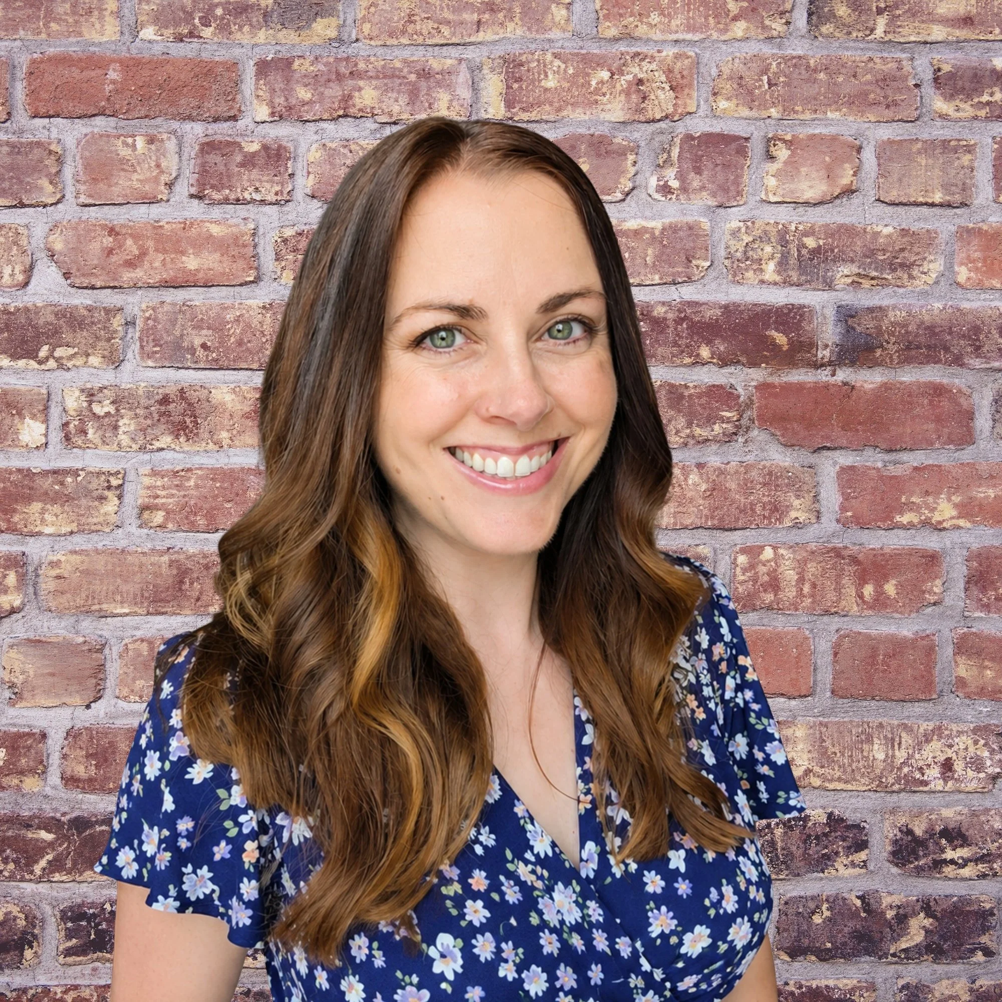 Close-up of a smiling woman with long brown hair and green eyes, wearing a blue floral dress, standing in front of a brick wall.