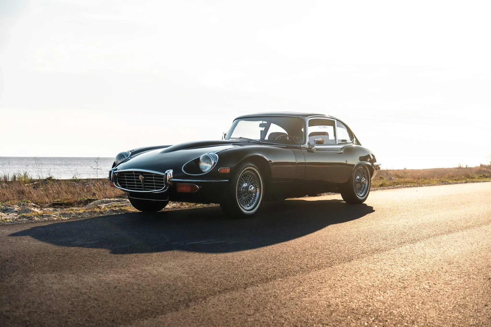 A Black Jaguar E-Type Coupe XKE parked on the side of a road near a beach with grass and a body of water in the background under a clear sky.