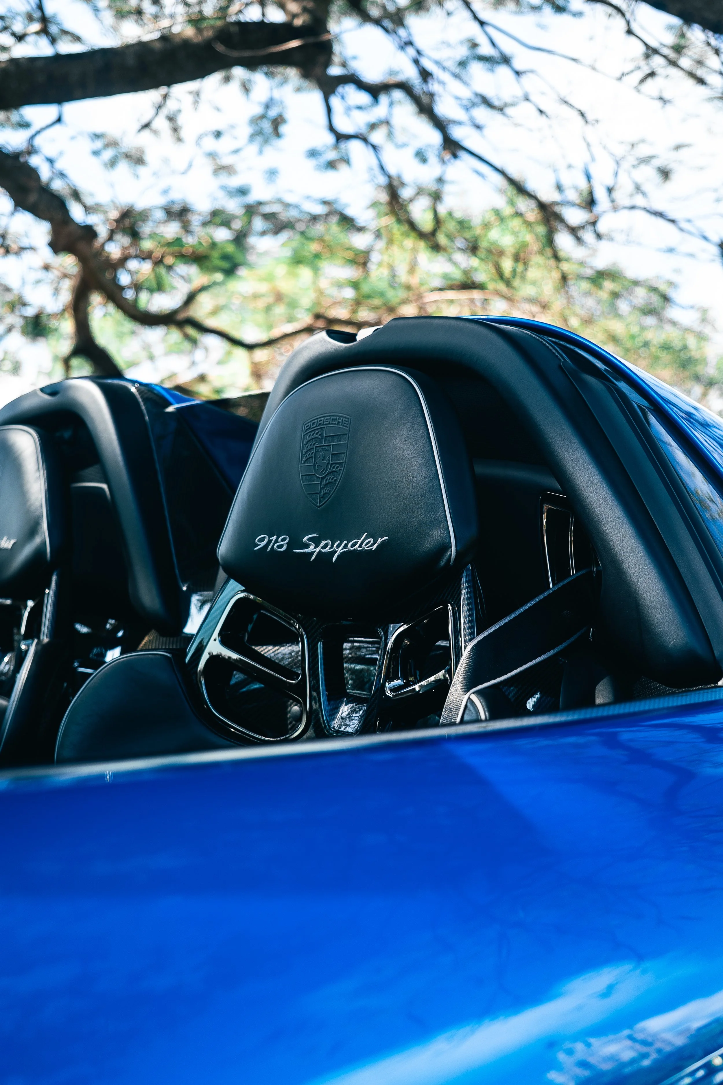 Close-up of a black headrest with the Porsche logo and '918 Spyder' embroidered on it, inside a blue Porsche 918 Spyder, with trees and sky visible outside.